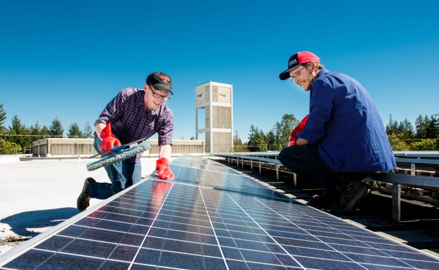 Two White male students sit smiling as they look on at a solar panel display on top of a building.