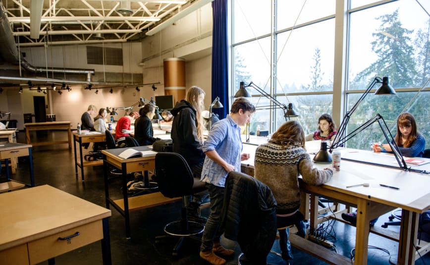 A group of students, most with the back to the camera, look on and work at architect tables.