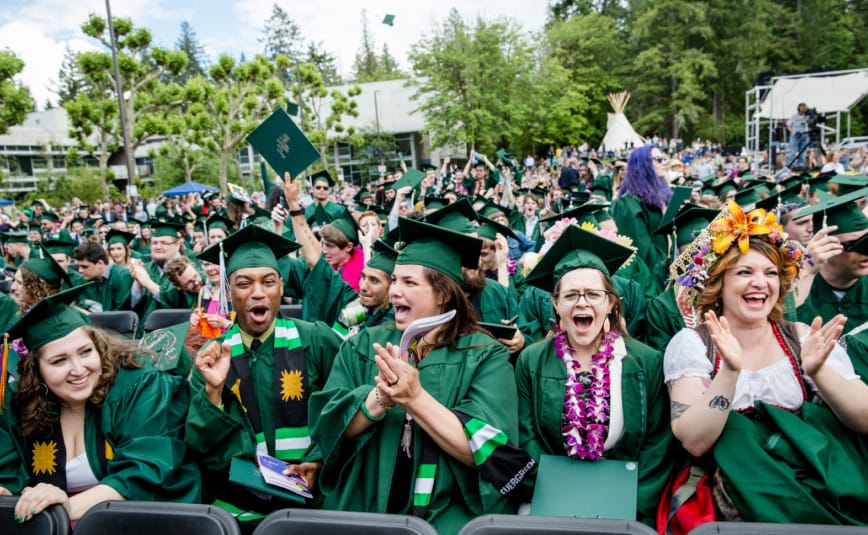 A group shot of many diverse students dressed in graduation regalia as they smile, cheer and laugh at the graduation ceremony.